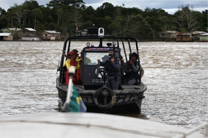 Police officers aboard a motorboat on a broad river, with houses and forest along the bank.