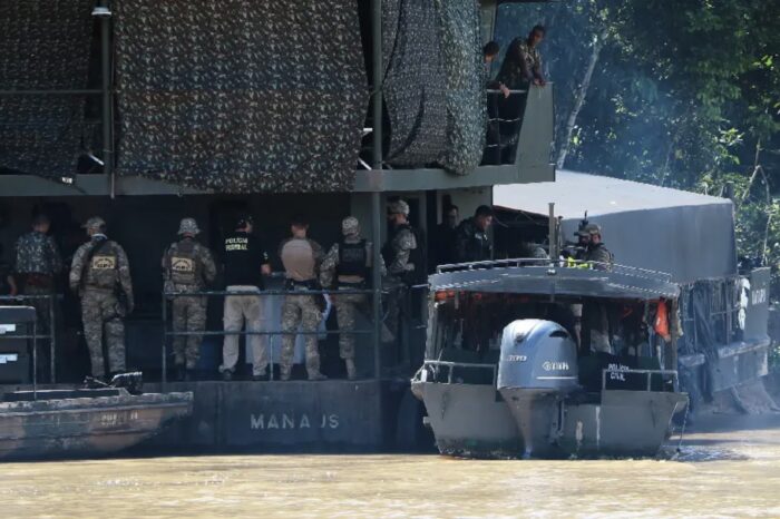 Uniformed personnel stand at a camouflaged floating base, with a police motorboat moored alongside.