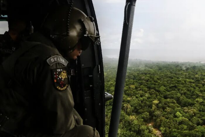 A helmeted soldier looks out through an open helicopter doorway above dense Amazon rainforest.