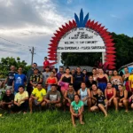 Residents of the Nova Esperança Indigenous community pose beneath a red and blue welcome arch, surrounded by greenery and community buildings.