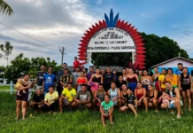 Residents of the Nova Esperança Indigenous community pose beneath a red and blue welcome arch, surrounded by greenery and community buildings.