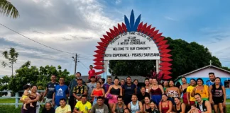 Residents of the Nova Esperança Indigenous community pose beneath a red and blue welcome arch, surrounded by greenery and community buildings.
