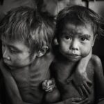 Brazil: The living dead Black-and-white photograph of two young Yanomami children sitting closely together, one looking down while the other looks towards the camera with an arm around the first child.